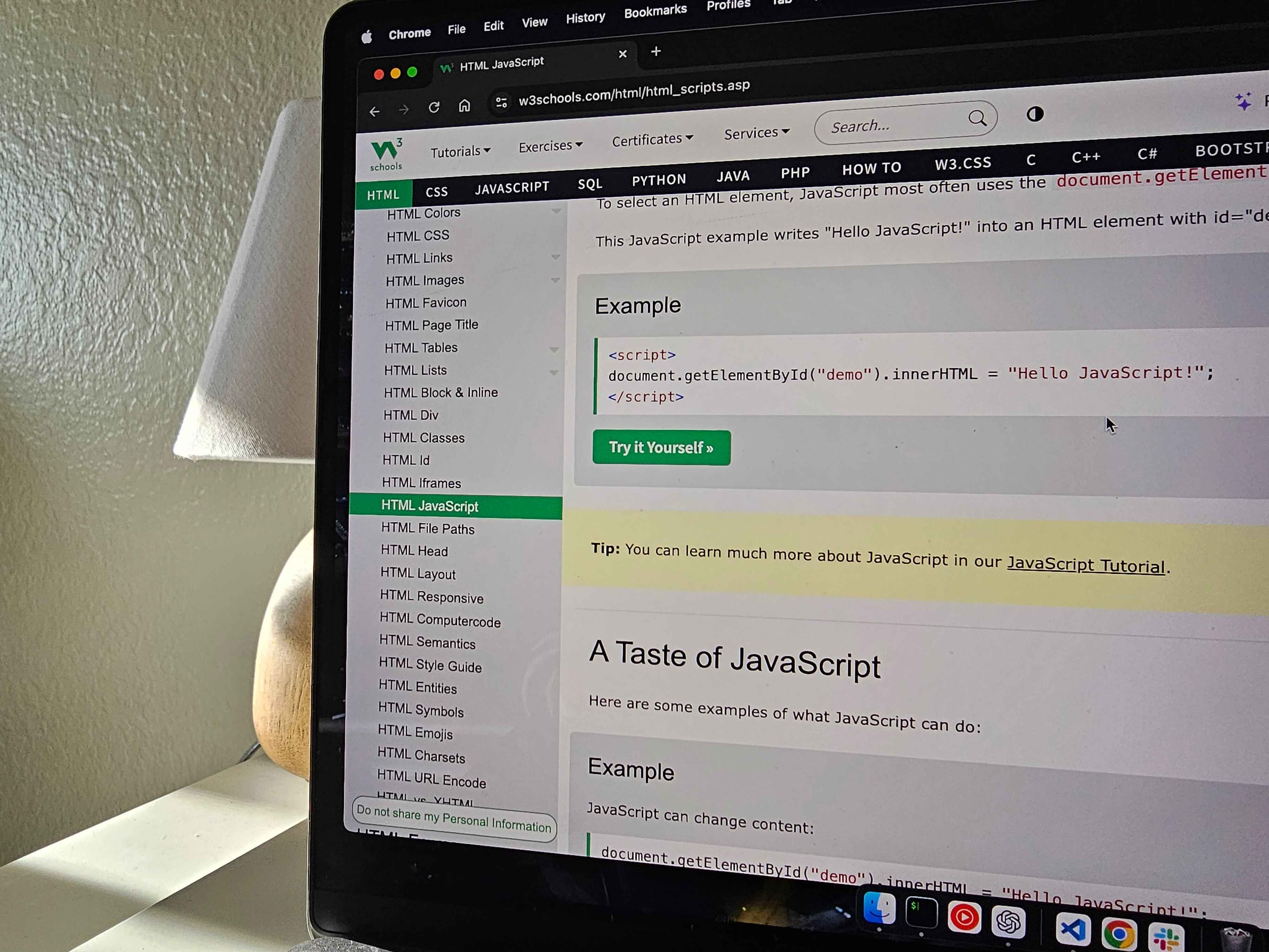 Hello JavaScript! An image of W3Schools pulled up on a laptop with a lamp behind the laptop, and sun shining on the desk